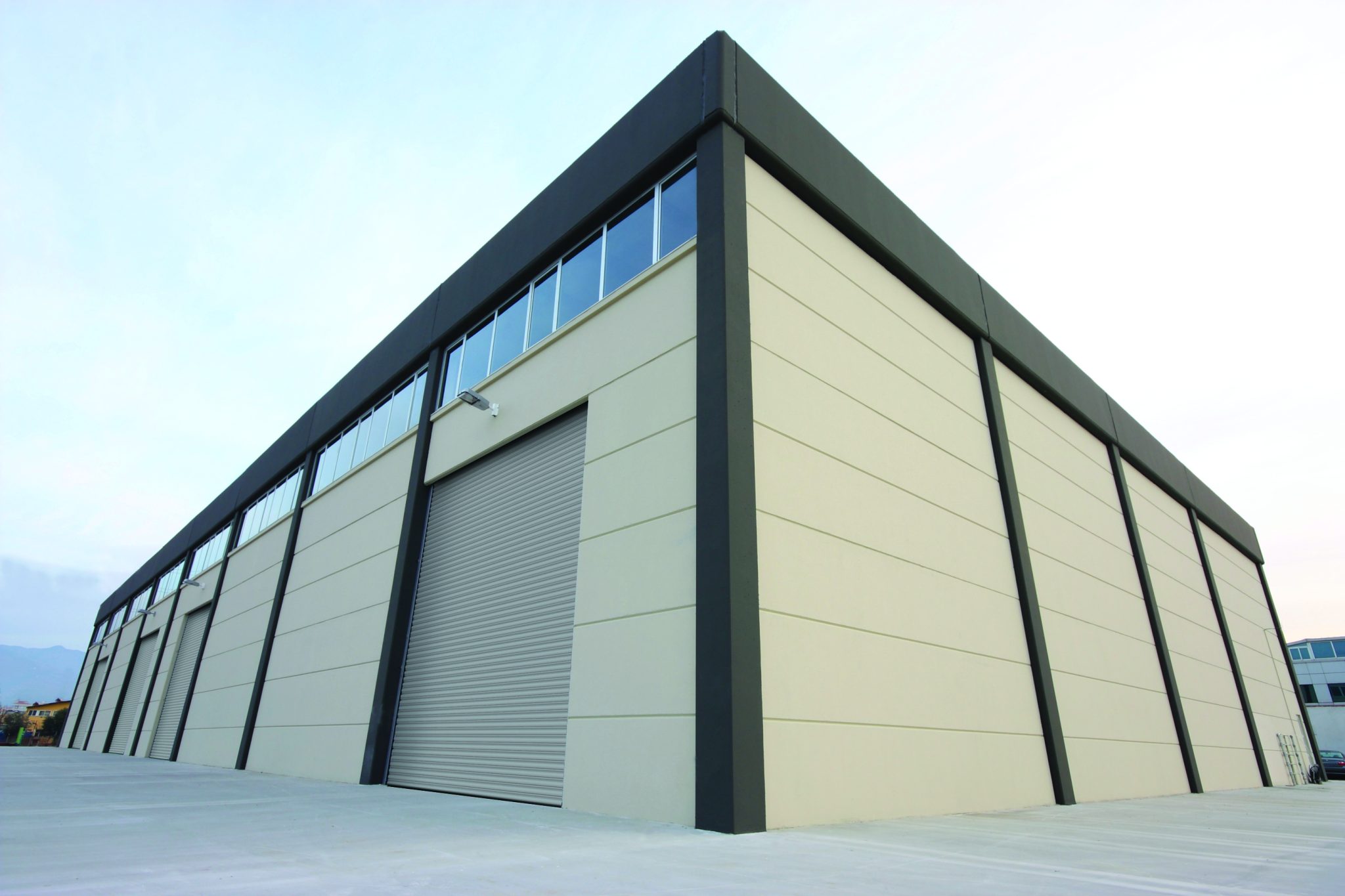 A modern, large warehouse building with beige walls and dark brown trim. The structure has multiple long windows near the roof and a central rolling door for vehicle entry. The area around the warehouse is paved with concrete, and the sky is clear.