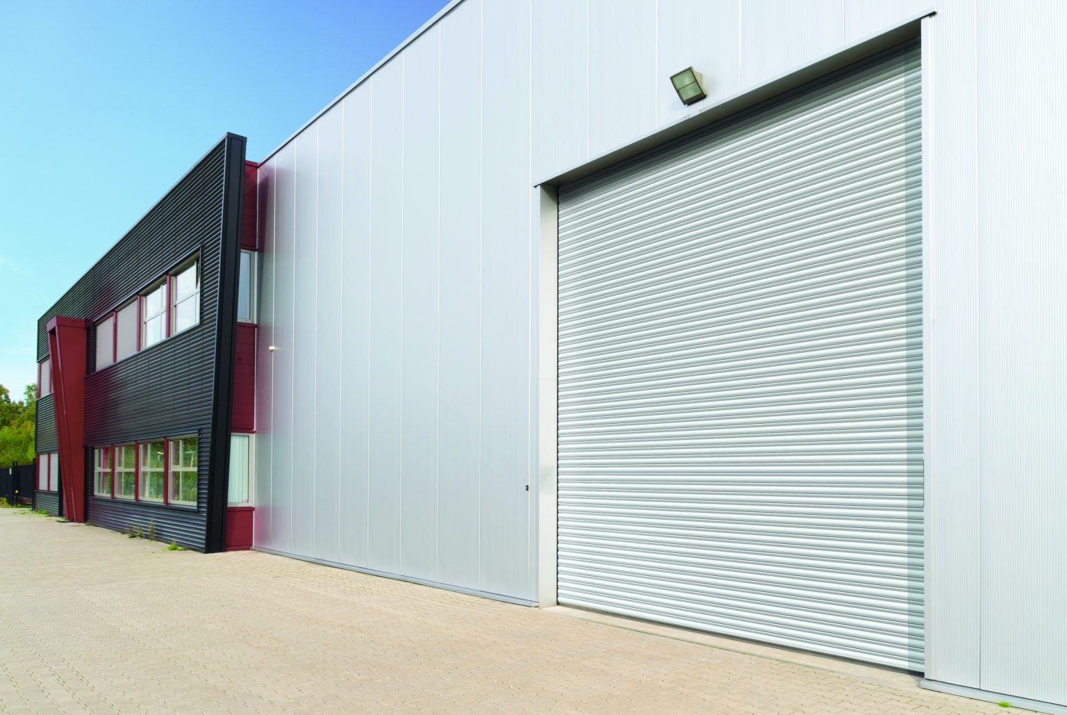 A modern industrial building with a large white roller door on the right side and gray metallic walls. The left side has black and red paneling with multiple windows. The building is surrounded by a paved area under a clear blue sky.