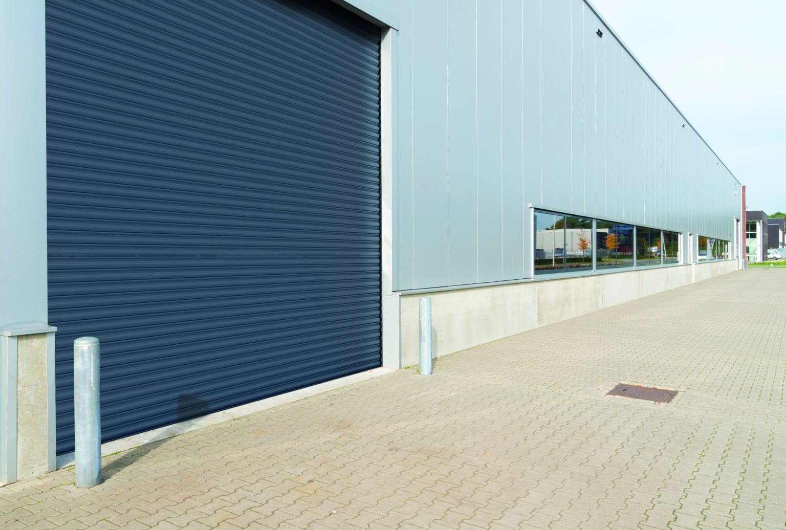 A modern industrial building featuring a large closed roller shutter door. The exterior has a combination of metallic panels and concrete base. There is a paved area in front and tall bollards stand near the entrance, with windows running along the length of the wall.