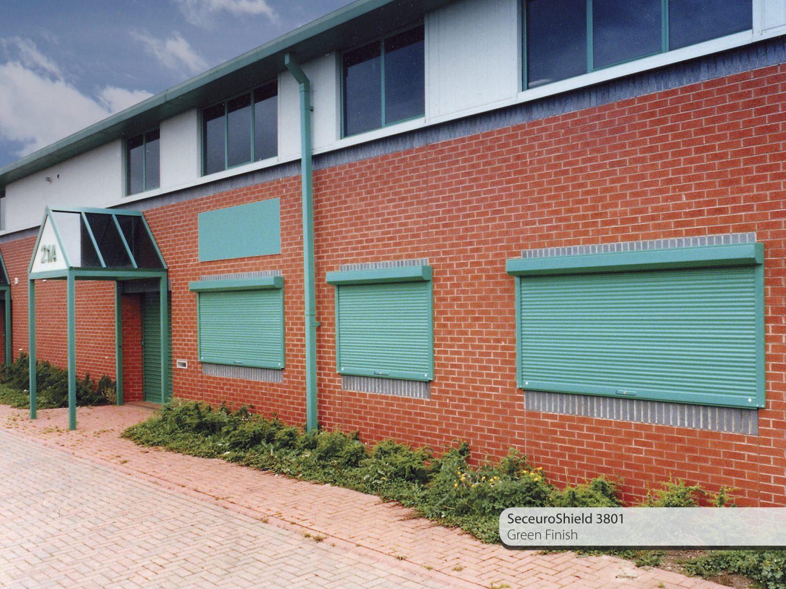 A brick building with green-colored security shutters on the windows and door. The building's entrance has a green awning with glass panels. Text in the image reads "SeceuroShield 3801 Green Finish." The building is surrounded by a paved walkway and some shrubbery.