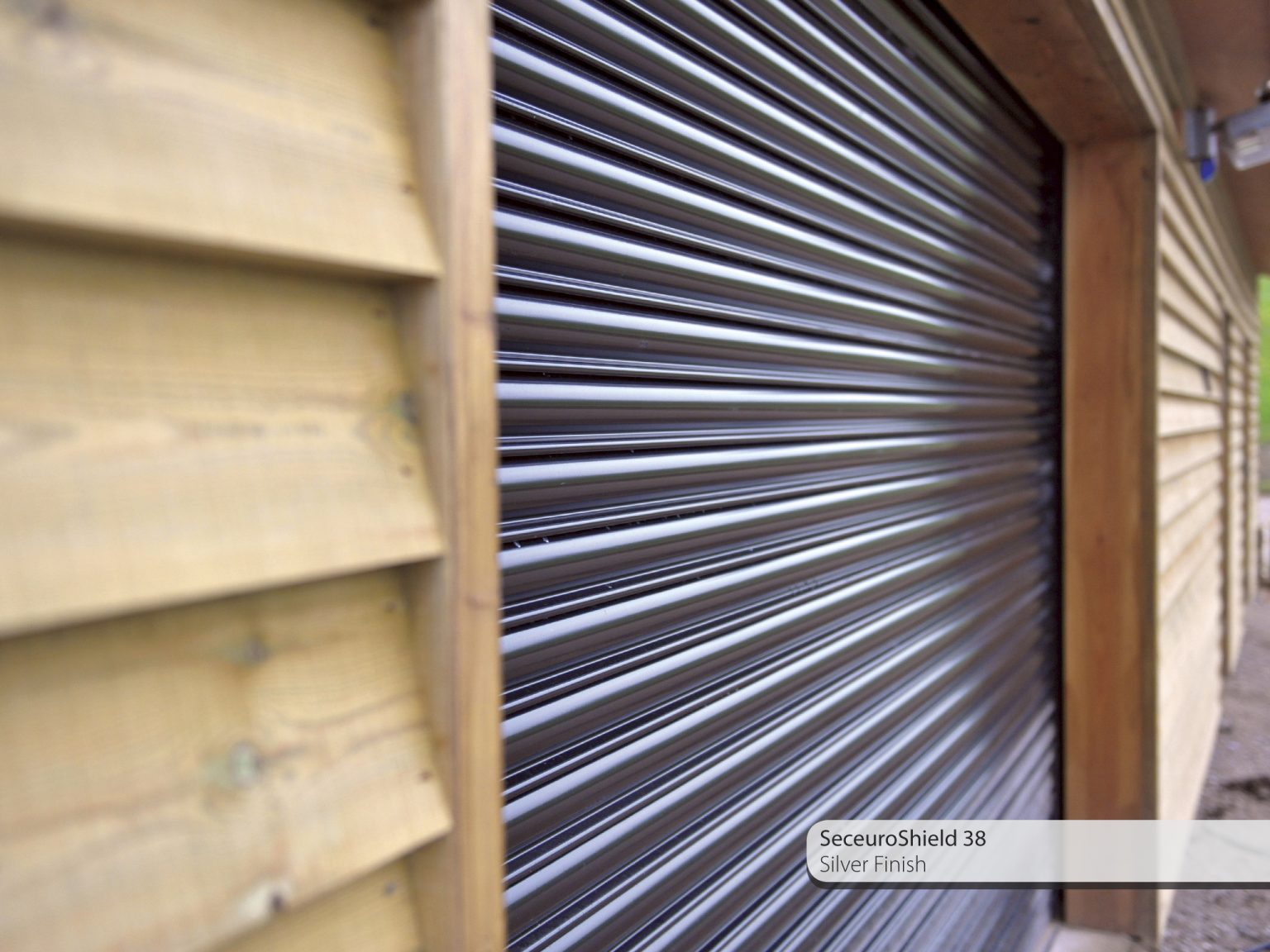 Close-up view of a metallic roller shutter door installed on a wooden exterior wall. The shutter has a silver finish, and text in the corner reads "SeceuroShield 38 Silver Finish.