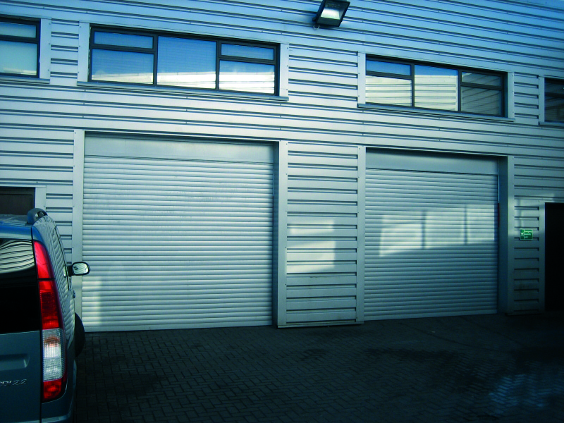 A blue vehicle is parked in front of a modern industrial building with two closed garage doors. The exterior features horizontal metal siding and large rectangular windows above the garages. The ground is paved with bricks.