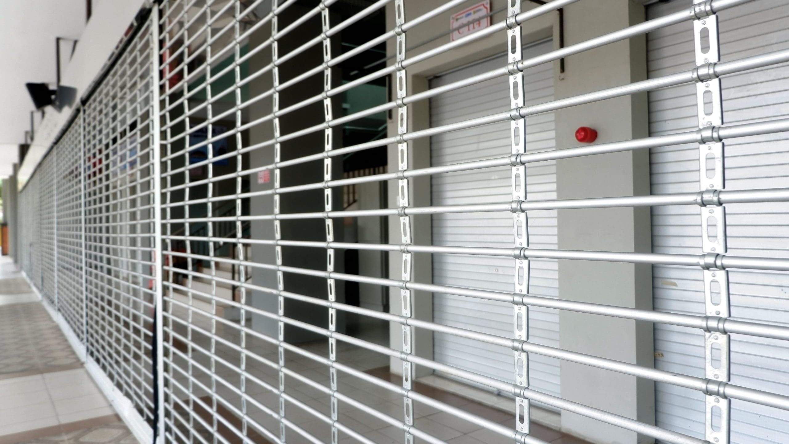 A view through a closed metal security grille, revealing shuttered storefronts inside a commercial building.