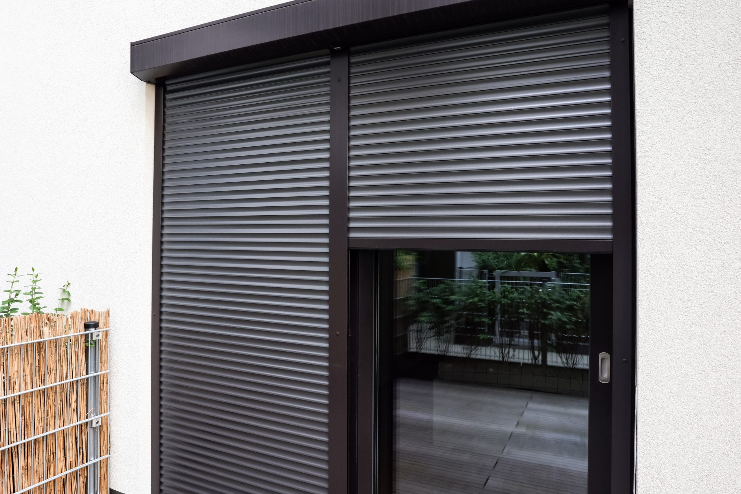 A modern building with a grey rolling shutter partially closed over a sliding glass door. The door reflects a balcony and greenery. A trellis with plants is positioned near the door on a white wall.