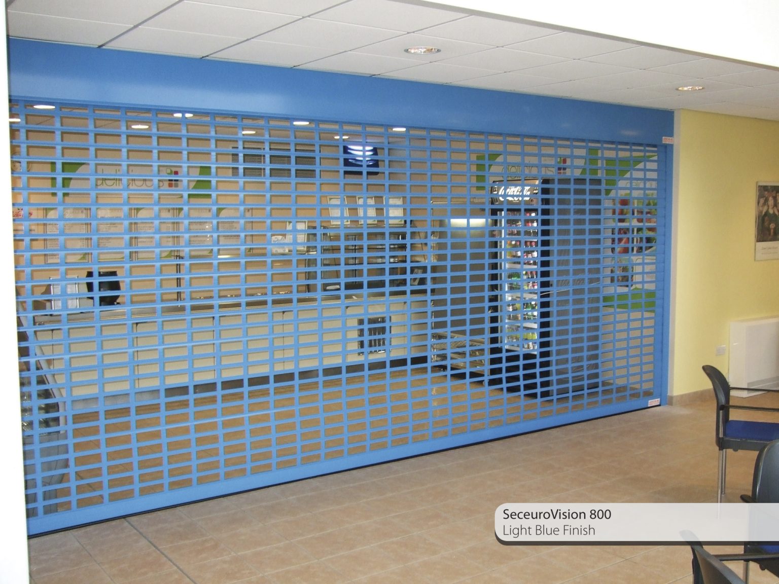 A metallic security shutter painted in a light blue finish is installed across an indoor commercial space. The shutter, labeled "SeceuroVision 800 Light Blue Finish," allows visibility of the well-lit interior, which includes a counter and an information area.