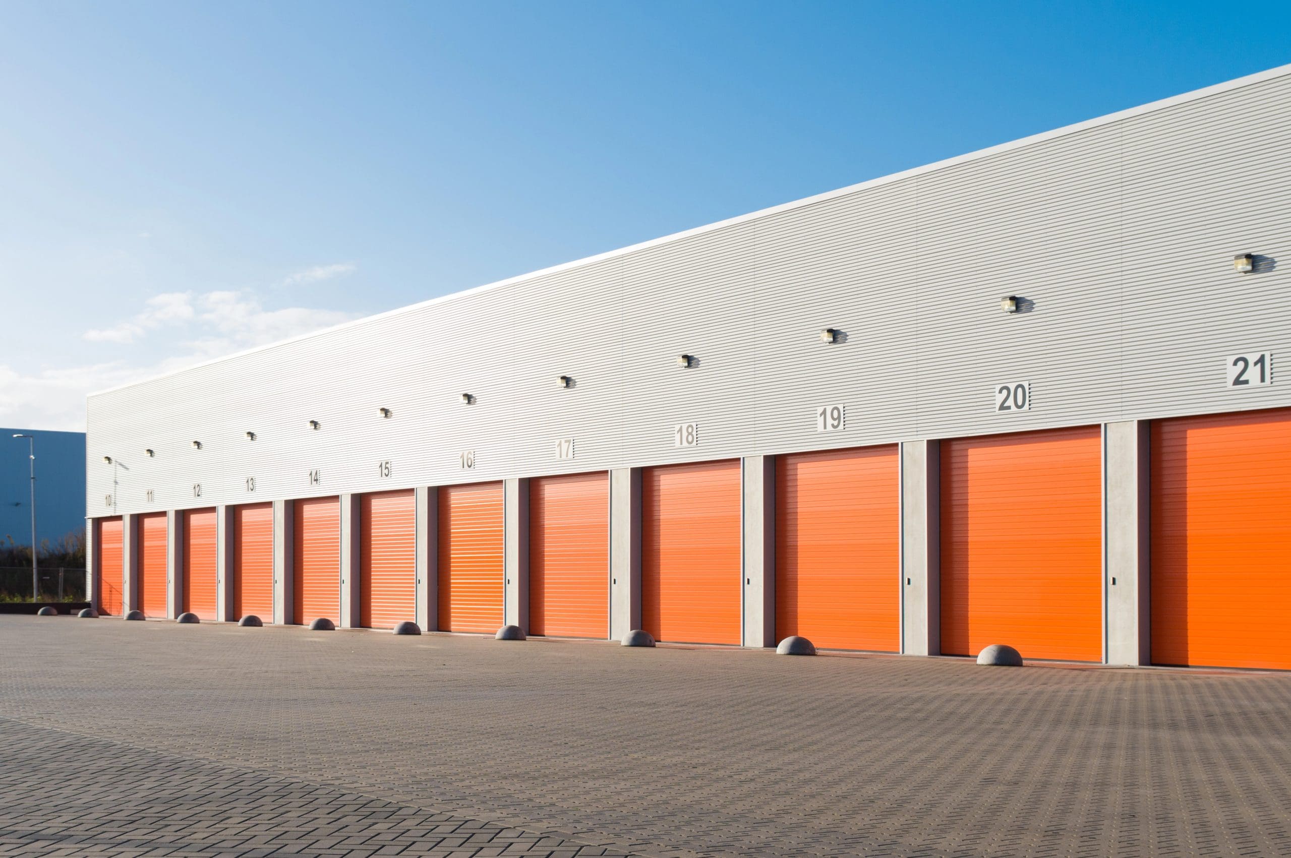 A row of large, orange storage unit doors lined up under a modern, white corrugated metal building. Each door is numbered sequentially from 15 to 21. The units are part of an outdoor storage facility. The sky is clear and blue.