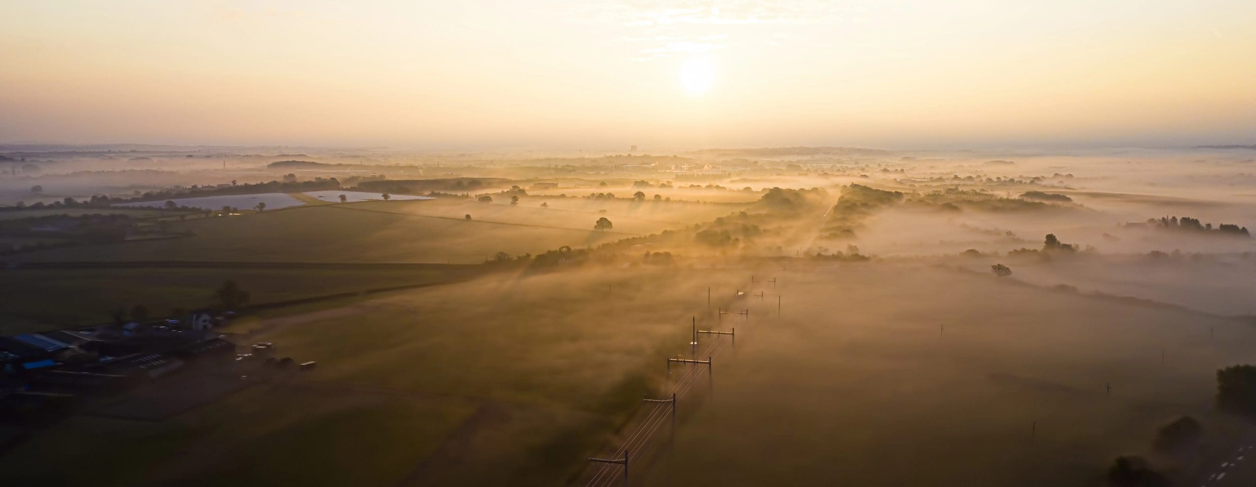 Aerial view of a tranquil, foggy landscape at sunrise. The soft, golden sunrise casts warm light over fields, dotted with trees. Misty, rolling hills are visible in the background, creating a serene and ethereal atmosphere.