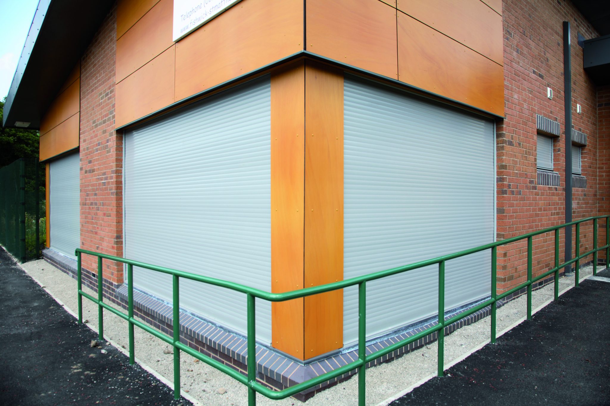 The image shows the corner of a modern building with light gray metal roller shutters covering the windows. The walls are made of a combination of brick and a wood-like material at the top. A green metal railing encloses the area in front of the building.