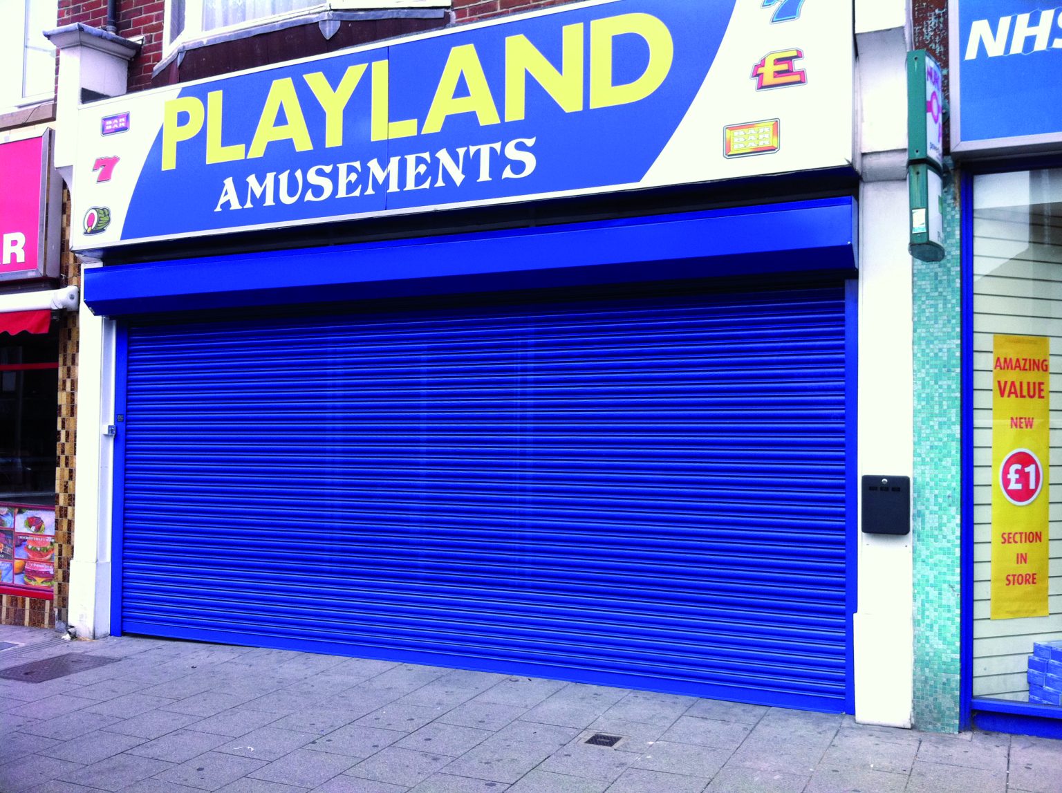 The image shows a storefront with a blue and yellow sign that reads "PLAYLAND AMUSEMENTS." The store's metal security shutter, also blue, is closed. Various small signs on the storefront include "£1" indicating price promotions. The sidewalk in front is empty.