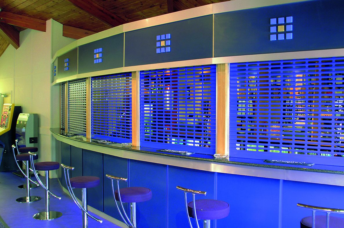 A modern bar with a blue-themed counter and stools. The counter features nine evenly spaced silver stools with round, blue seats. The backdrop consists of metallic, grid-patterned shutters, each section outlined with blue panels.