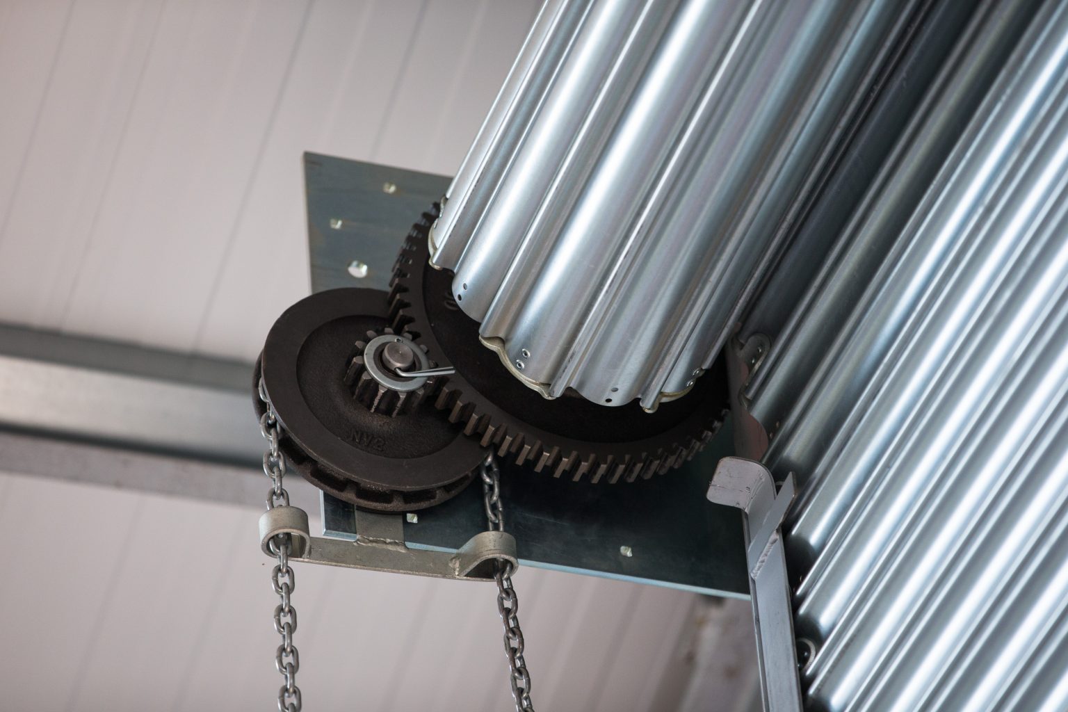 A close-up of a metal roller door mechanism. Two large gears are connected by a chain, with cylindrical sections of the door coiled neatly above. The structure is mounted on a metal frame attached to a ceiling.