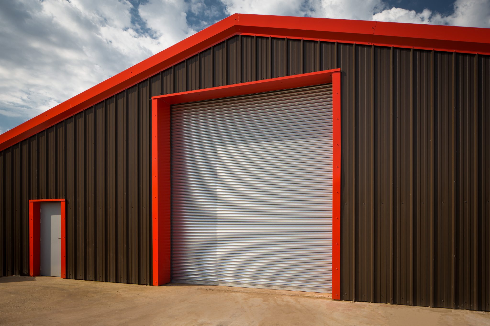 A large industrial building with a corrugated metal exterior. It features a high, wide closed garage door and a smaller adjacent door, both framed in red. The sky above is partly cloudy, casting soft shadows on the building.