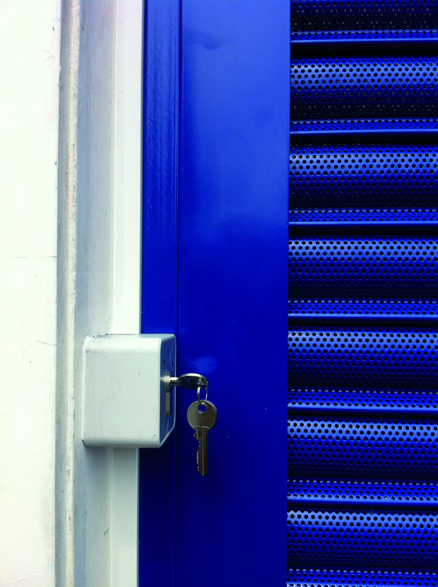 A close-up of a bright blue metal door with a perforated design on the right. A silver lock box is mounted on the left side of the doorframe, with a set of keys inserted into it. The background is a white wall.